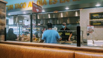 Marcos Carbajal, dueño del restaurante Carnitas Uruapan, toma la orden de un cliente detrás de un plástico protector mientras su empleada le entrega a otra cliente su comida para llevar. (Irene Tostado / La Raza)
